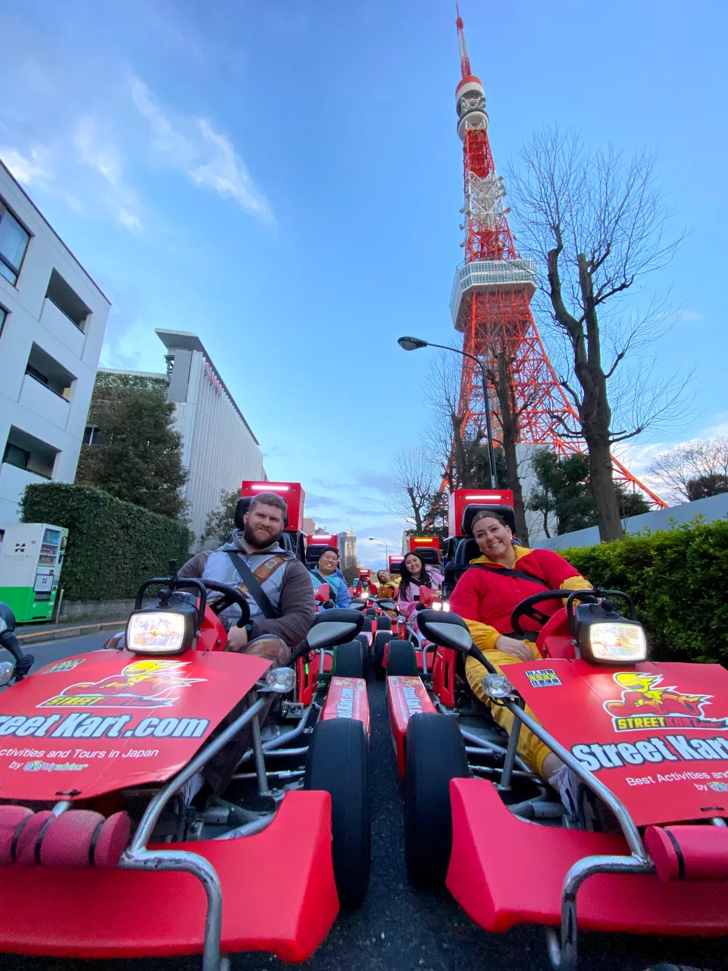 People ride red street karts on a city street with Tokyo Tower visible in the background under a blue sky, wearing seatbelts and smiling guardrails nearby.
