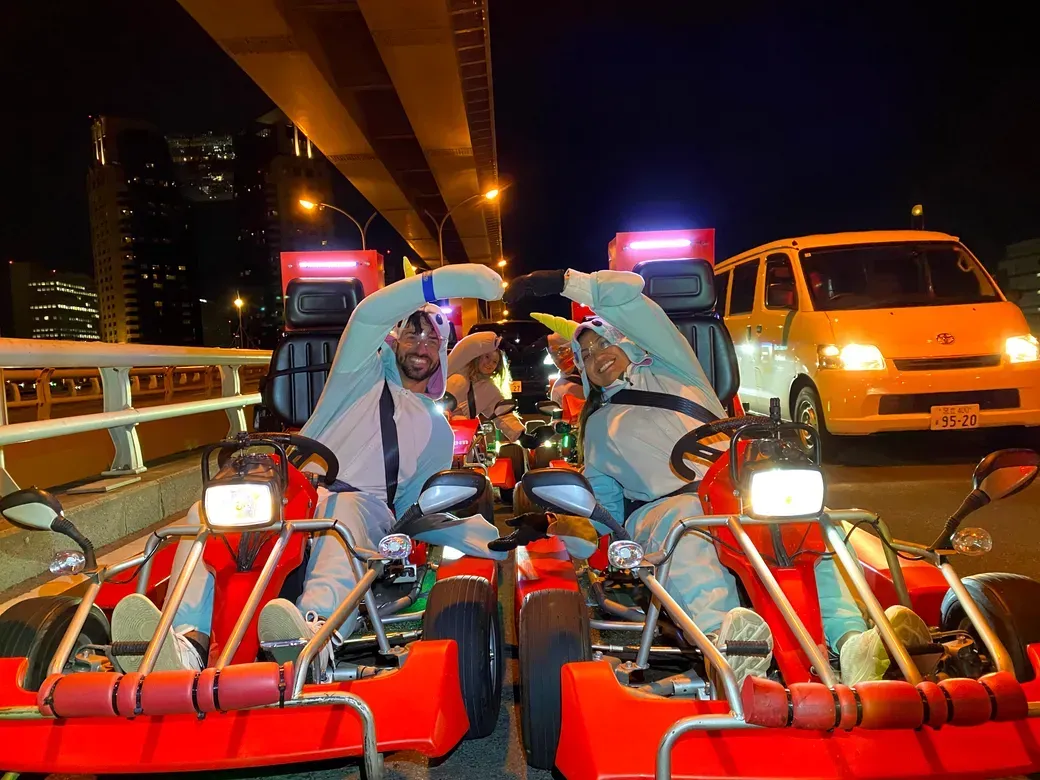 Two people in white outfits in bright red go-karts on a city street at night, forming a heart shape with their arms.