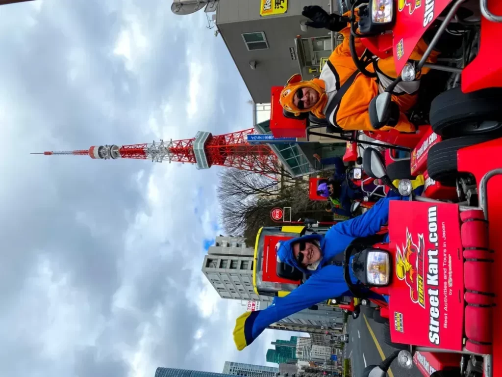 A group of people in bright costumes ride red go-karts on a city street, with Tokyo Tower visible in the background.