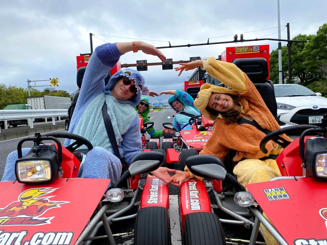 Group of friends in colorful onesies posing in red go-karts on a racetrack, forming a heart shape with their arms.